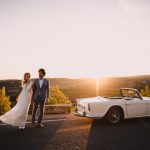 Bride and Groom by white wedding car in sunset
