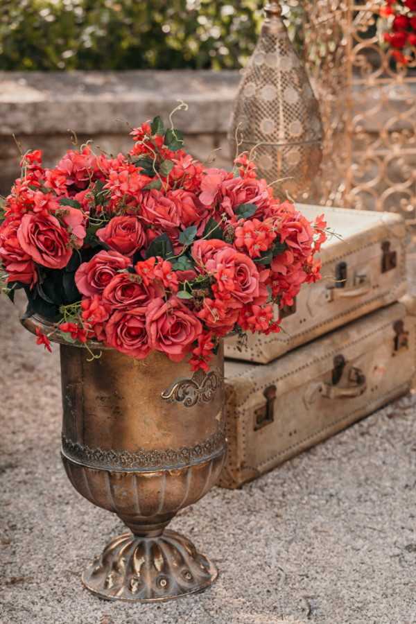 Red flowers fill antique brass vase next to vintage luggage