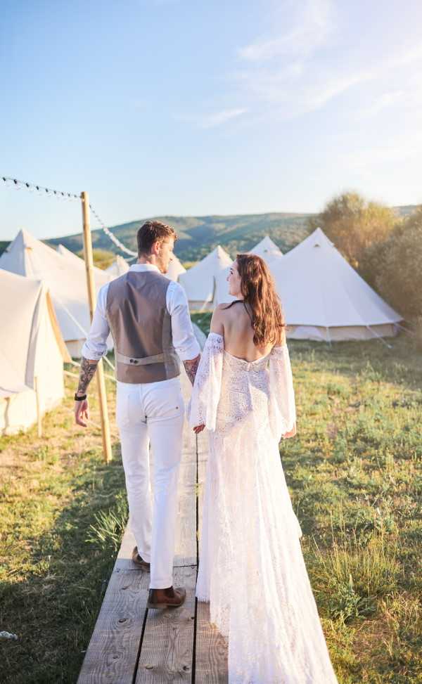 Bride and groom walk hand in hand in the afternoon sun by row of cotton tents for wedding guests