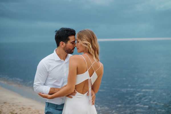 Groom in blue suit pants and bride in white silk open back gown by the sea