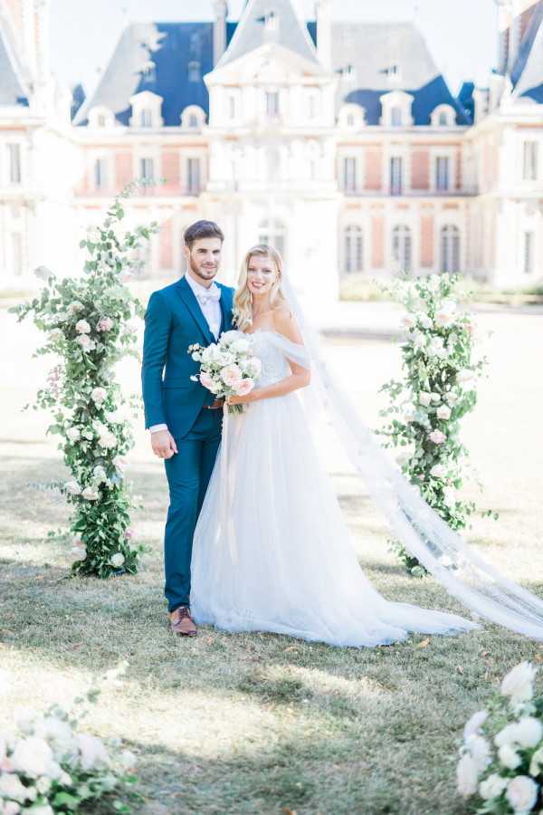 Bride and groom pose in front of white rose arbour outside Chateau de Baronville, France