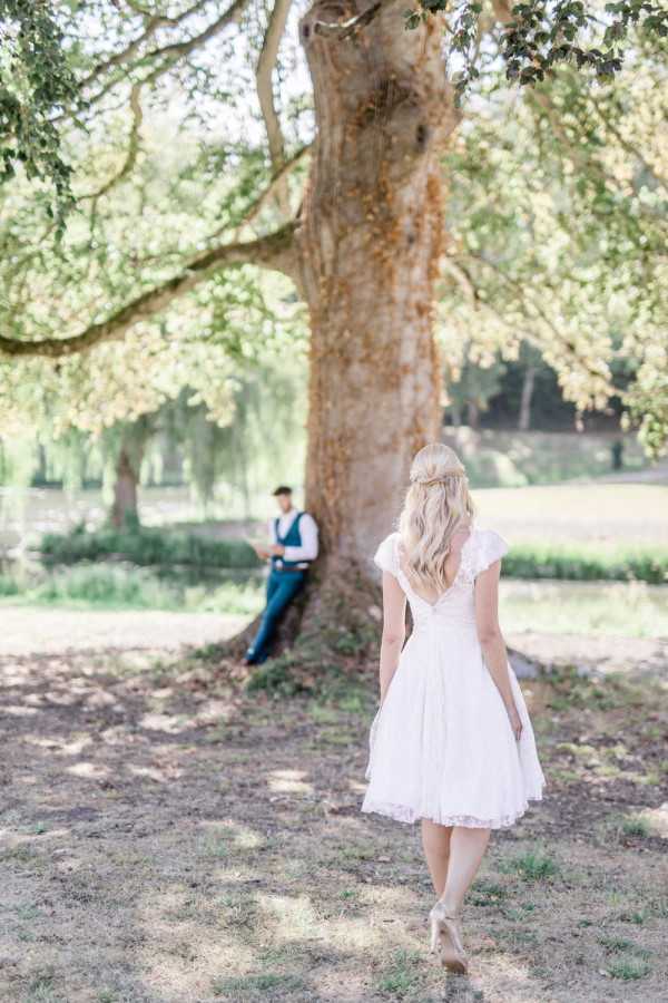 Bride in short white dress approaches groom in blue waistcoat reading under tree