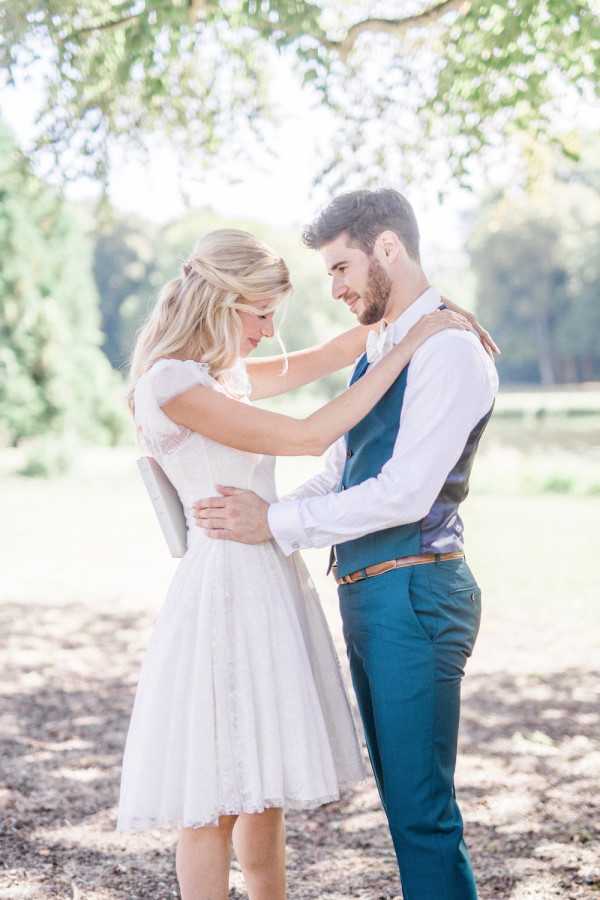 Bride in short white dress and groom in blue waistcoat embrace outdoors