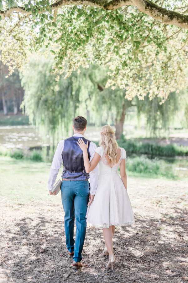 Bride and groom walk away from camera towards willow trees outdoors holding books