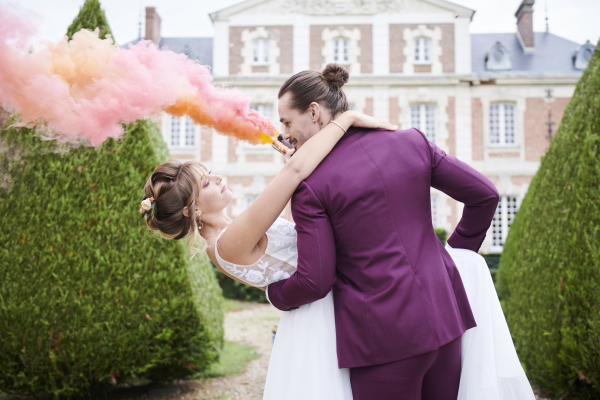 Groom in purple suit tips his bride back and purple and orange smoke blooms behind them
