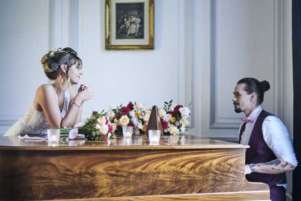 groom in purple waistcoast plays piano to his bride who leans on the piano