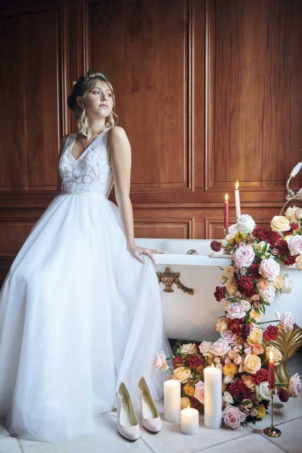 bride sits on edge of bathtub surrounded by flowers and candles in white lace gown