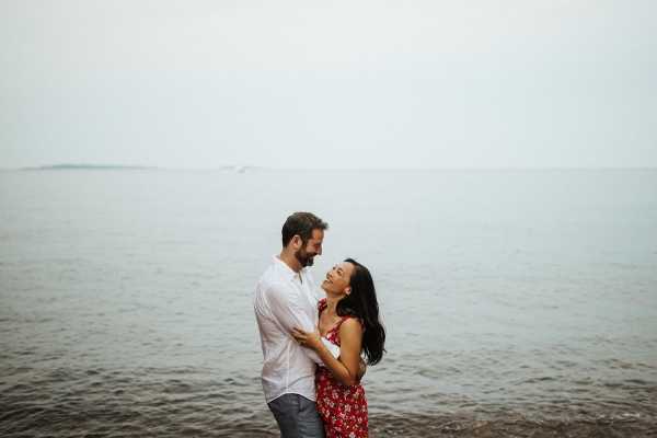 Man in white shirt and woman in red dress embrace by waters edge