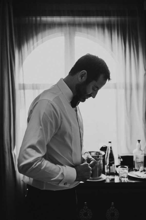 Black and white image of groom getting ready and framed by window