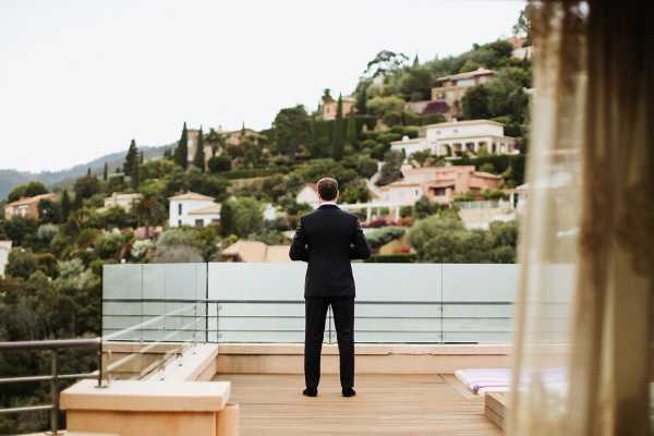 Groom in suit waiting for bride on balcony of hotel with back turned to camera