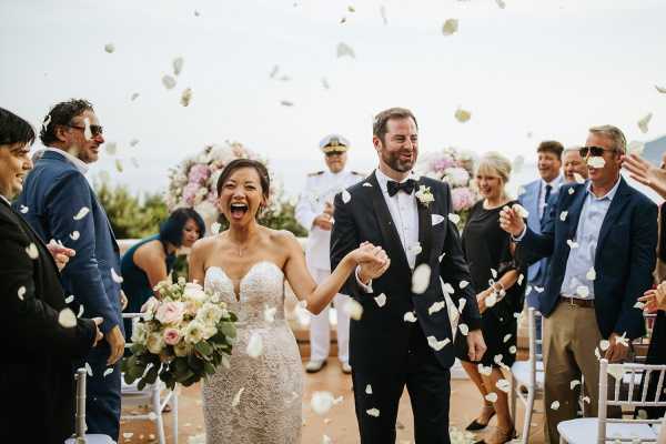 Just married couple smile brightly as wedding guests throw white confetti over them