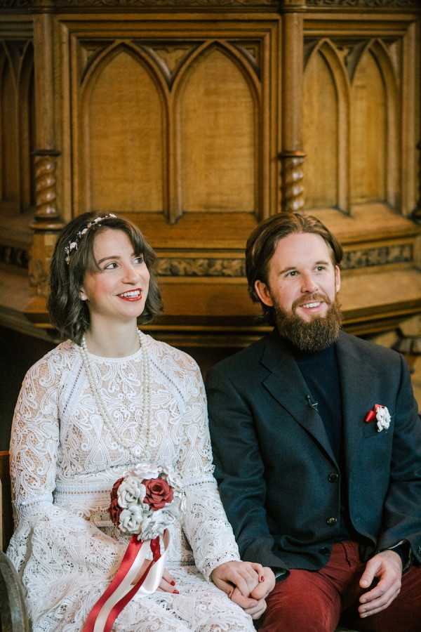 Bride and groom sit next to each other wearing black suit with black turtle neck and lace dress with pearl necklace and recycled paper bouquet