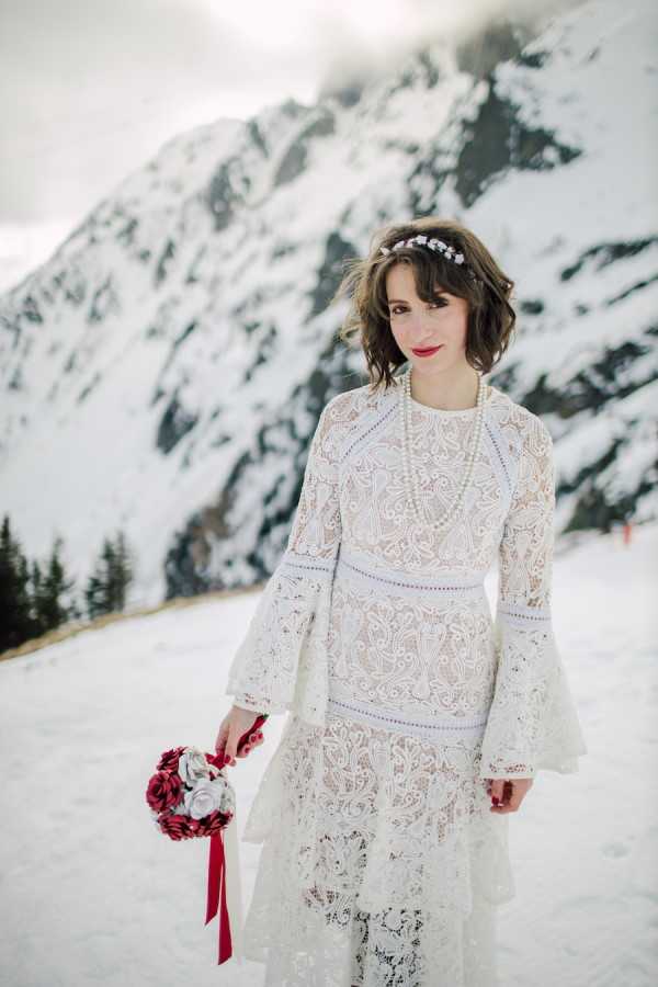 Bride in sustainable lace wedding dress with long bell sleeves and red lipstick stands in the snow in the French Alps