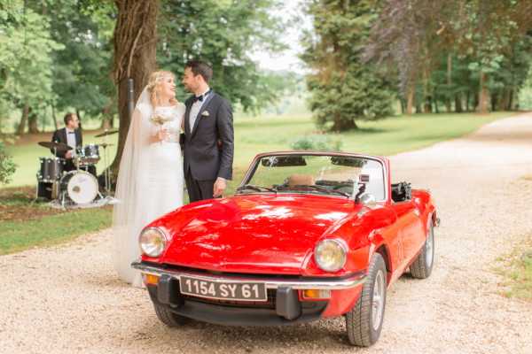 Bride and groom stand next to red vintage convertible car on the drive of Chateau d'Azy