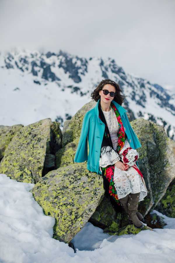 Bride in wedding dress, green woollen coat and grey boots with red lipstick sits on rocks on French Alps mountain top