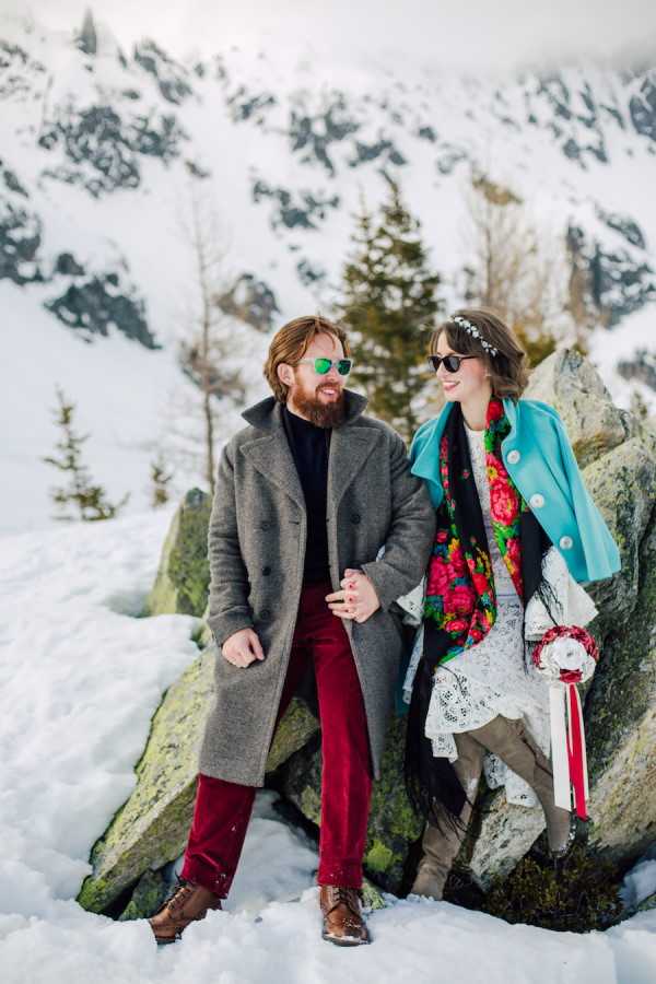 Groom in grey woollen coat ad red pants with sunglasses looks and smiles at bride in lace dress, green woollen coat and red scarf wearing sunglasses