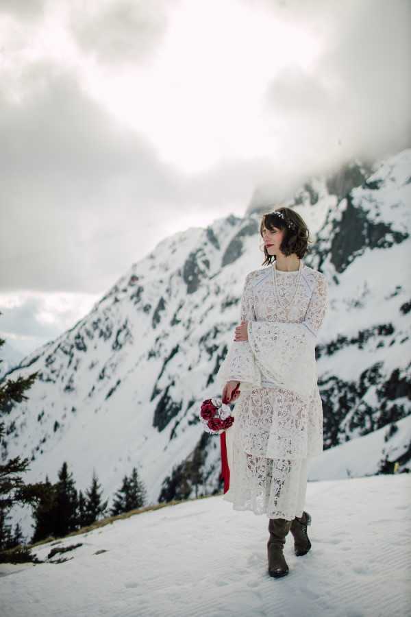 Bride in sustainable lace wedding dress with long bell sleeves and red lipstick stands in the snow in the French Alps for her Zero waste wedding