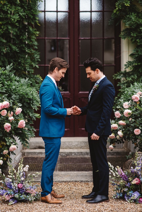 gay couple wearing blue and black suits, get married surrounded by flowers in front of tall brown french doors