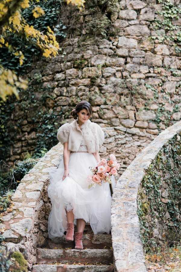 bride descends stone garden steps in angora jacket and pink suede boots under wedding dress