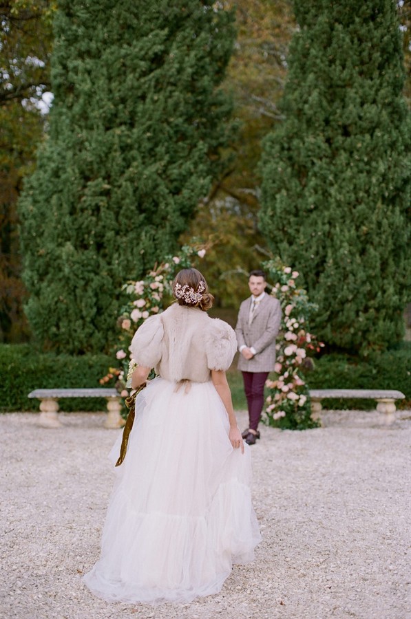 bride walks toward groom standing at floral archway in garden