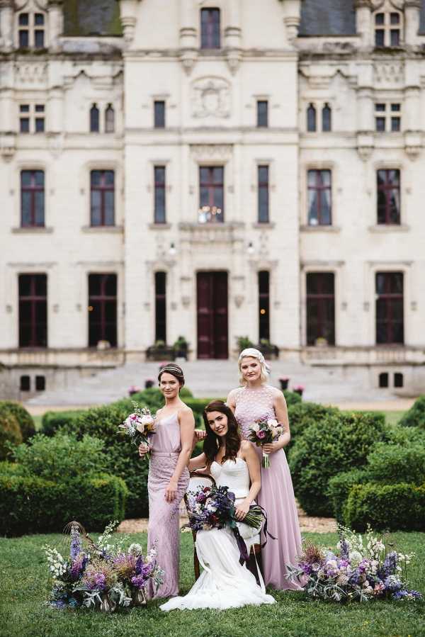 bride seated and 2 bridesmaids standing next to her in purple in front of chateau challain surrounded by purple flowers