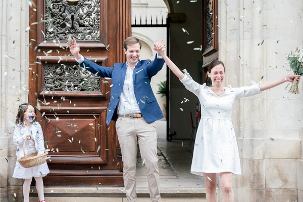 Bride and groom leave town hall with arms in air showered by confetti