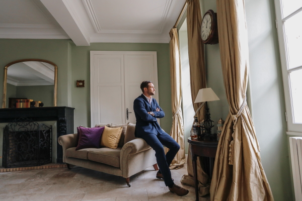 Groom waits before wedding ceremony in green walled room of Chateau Lacanaud sitting on the arm of a lounge