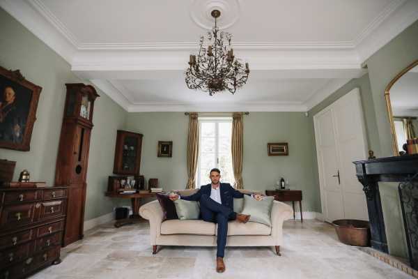 Groom in navy blue suit sits with one leg cross and arms outstretched on cream lounge in green room of Chateau Lacanaud in France