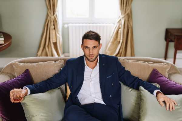 Groom looks into camera with arms outstretched on cream couch framed by gold silk curtains