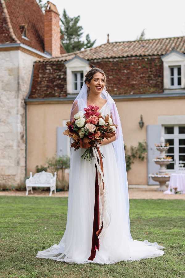 Bride stands outside Chateau Lacanaud holding burgundy and white bouquet and smiles towards her groom