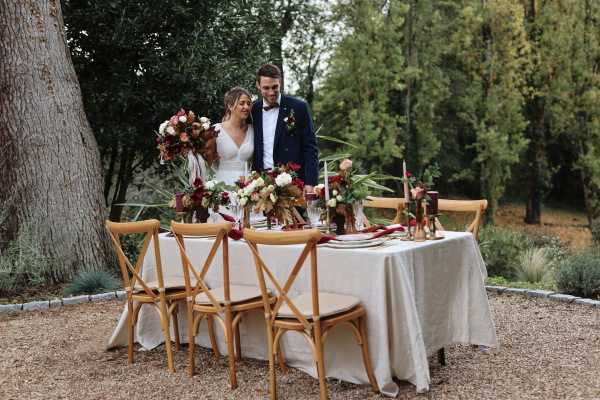 bride and groom stand behind their outdoor wedding table adorned in white table cloth and burgundy and pink table decorations
