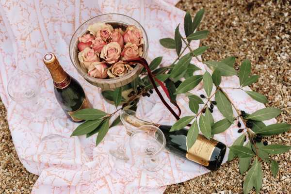 flatlay image of a bowl of pink roses, 2 bottles of champagne, a pink picnic rug and eucalyptus leaves