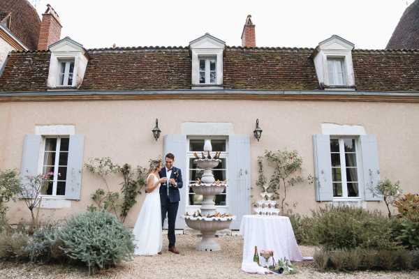 standing outside chateau lacanaud in dordogne france, bride and groom toast their wedding day next to fountain of champagne bottles