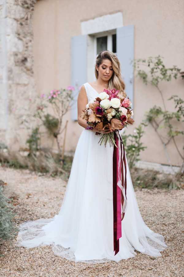 bride stands outside chateau lacanaud in france wearing white flowing bridal gown and holds burgundy large bouquet with long burgundy ribbons