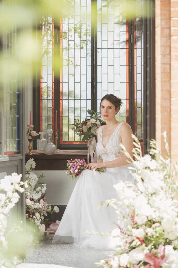 bride seated holding pink posie in front of dark wood bay window