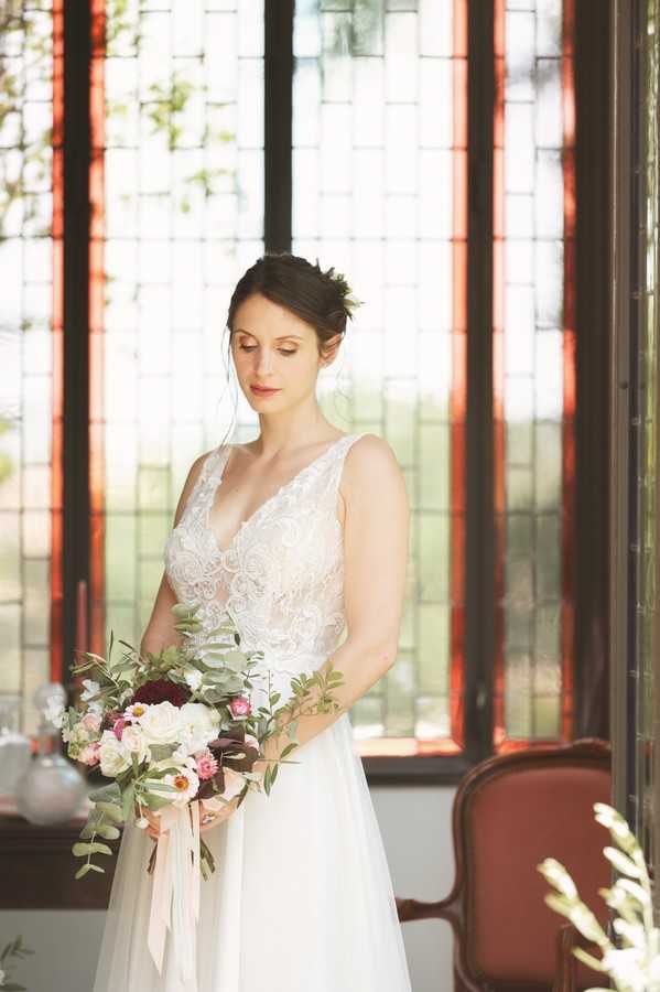 bride looks down and holds bouquet in front of dark wood bay window