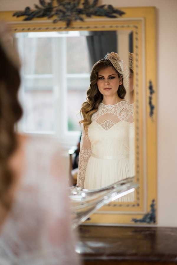 bride looks into framed mirror wearing long sleeved lace wedding dress and fascinator with netting over her face