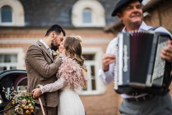 bride in pink feather boa and groom in brown tweed suit kiss next to accordion player