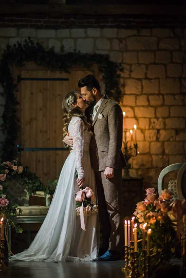 Newlywed couple kiss in candlelit rustic room surrounded by flowers