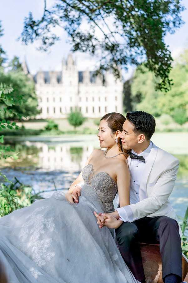 asian bride in dusty blue gown and her husband in white suit jacket sit under a tree with chateau challain visible in the background