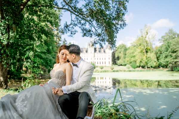 asian bride and groom sit under a tree in front of a lake with chateau challain visible in the background