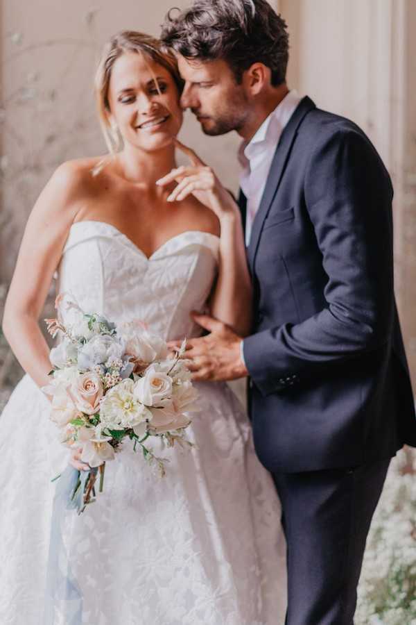 Bride and groom pose for the camera and look down at pastel bouquet