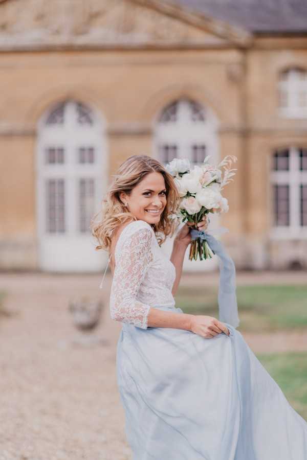 Bride in pastel blue skirt with white lace top holds bouquet wrapped in pale blue fabric to match her skirt