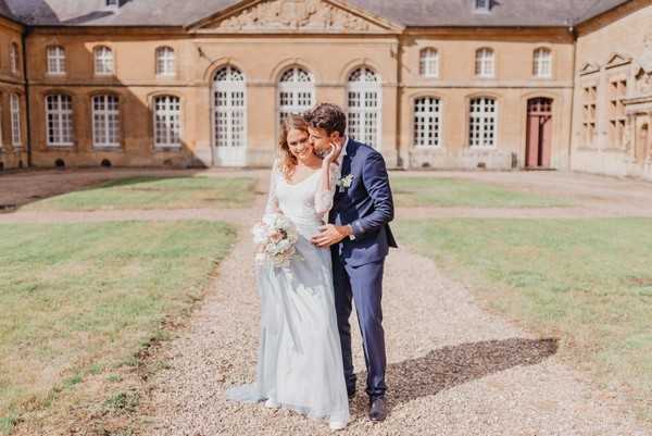 Bride in pale blue skirt and groom in midnight blue suit stand outside French Chateau