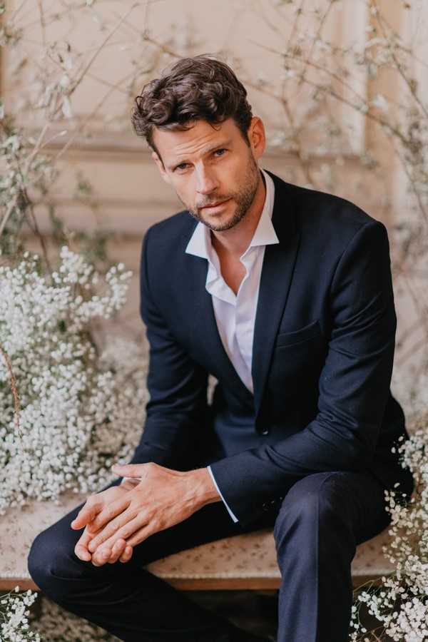 Groom in midnight blue suit and white unbuttoned shirt surrounded by white flowers