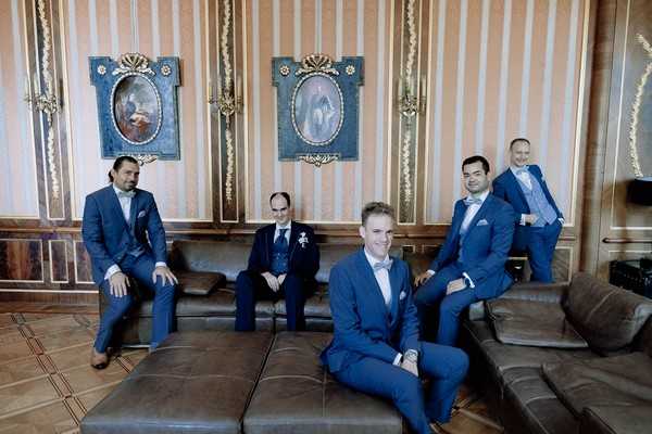 groom and groomsmen posed on brown leather lounges in front of ornate wallpaper and artworks