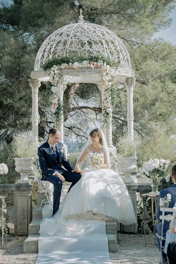 bride and groom sit in front of white ornate garden rotunda holding hands 