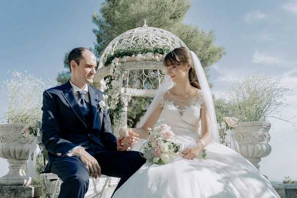 bride and groom seated hand in hand in front of garden rotunda decorated with pink roses