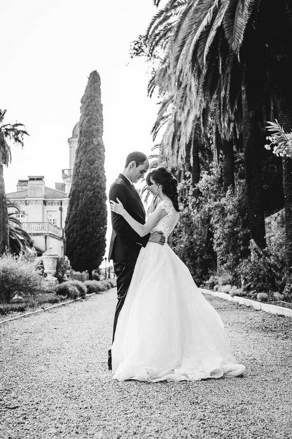 black and white image of bride and groom embracing on pebble garden path surrounded by trees and palms