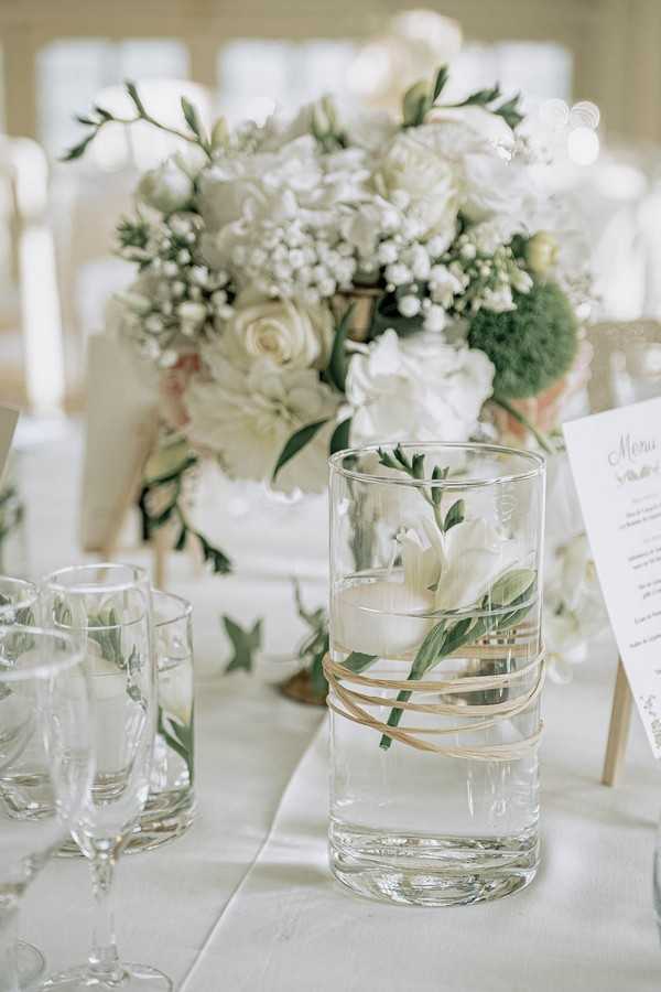 floral decorations for wedding table including vase of white flowers and a glass of water with small flower trimmings in it